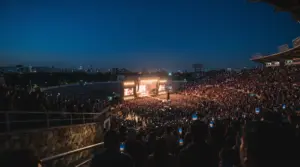 Estadio lleno al anochecer durante un concierto, con el escenario iluminado, pantallas y celulares encendidos entre el público